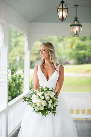 A bride in a white wedding dress stands on a covered porch holding a lush white and green floral bouquet. ive and oak wedding photos