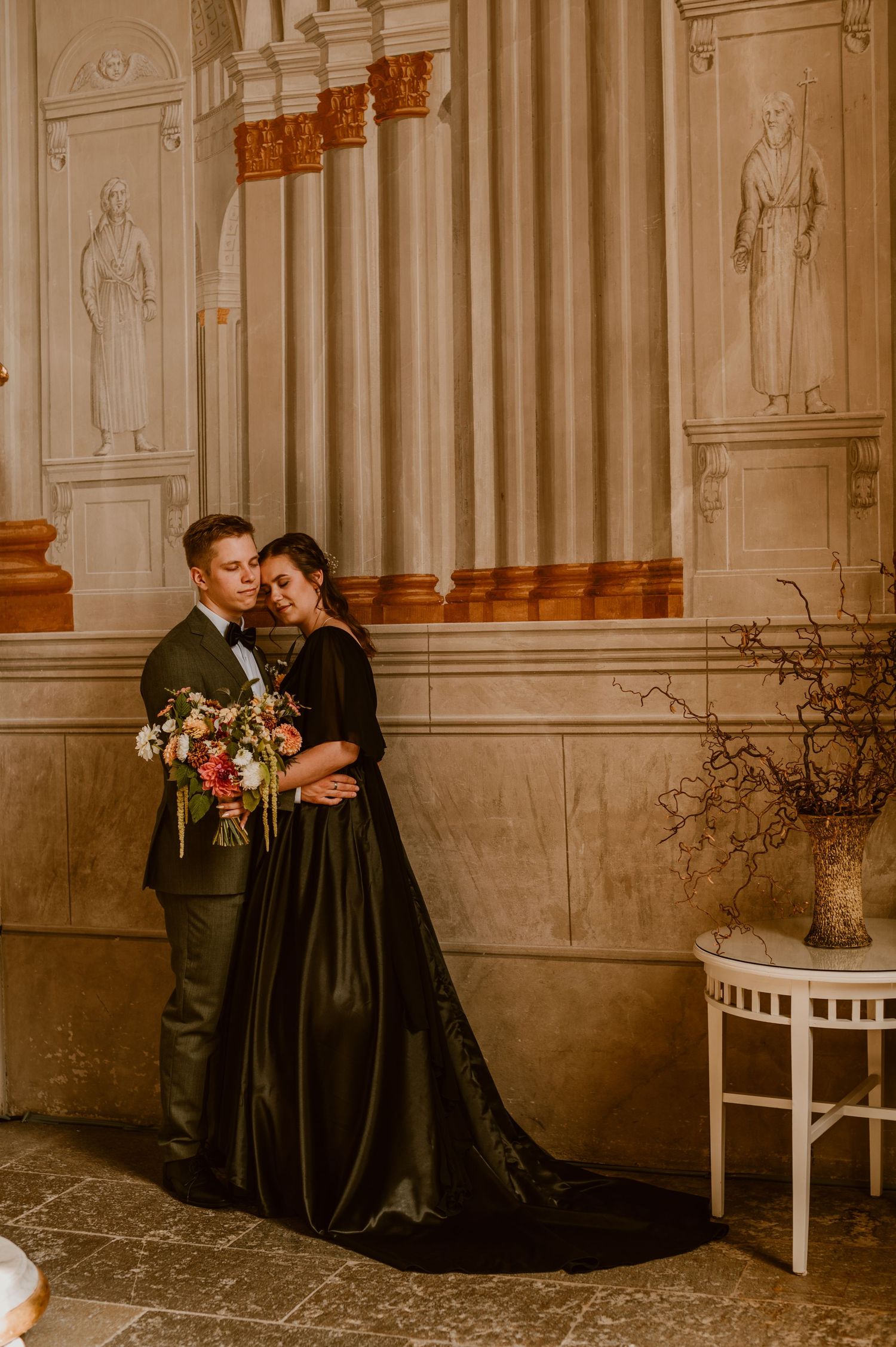 A couple in elegant black attire share an intimate moment in a grand room with ornate white walls and classical decor.