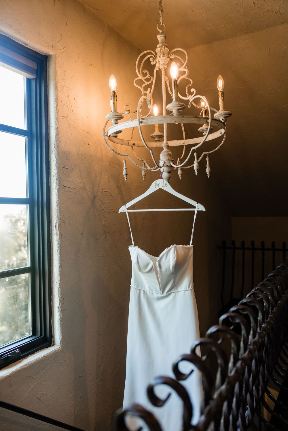 Elegant white wedding dress hanging from ornate chandelier near window in rustic setting.