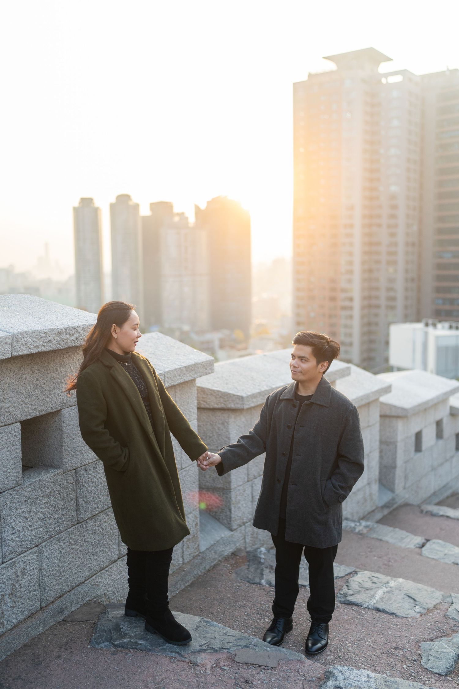 Two figures stand on a rooftop at sunset with city buildings visible in the background.