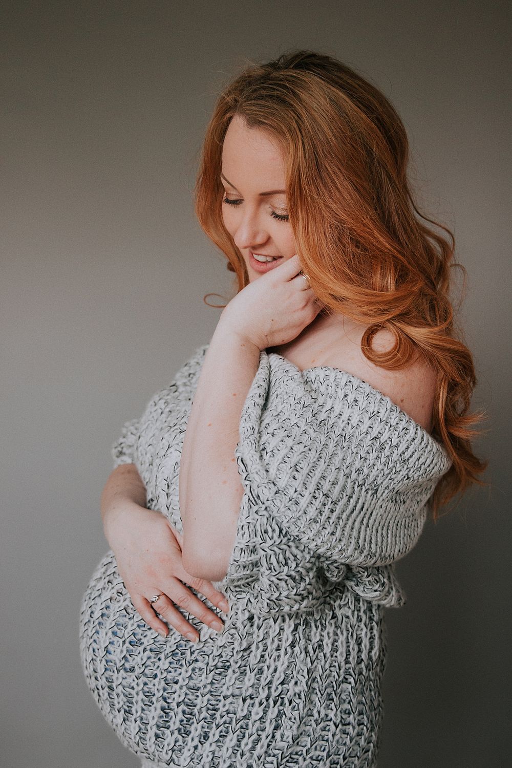 Expectant mother in gray knit sweater dress poses tenderly during maternity photoshoot with soft lighting.