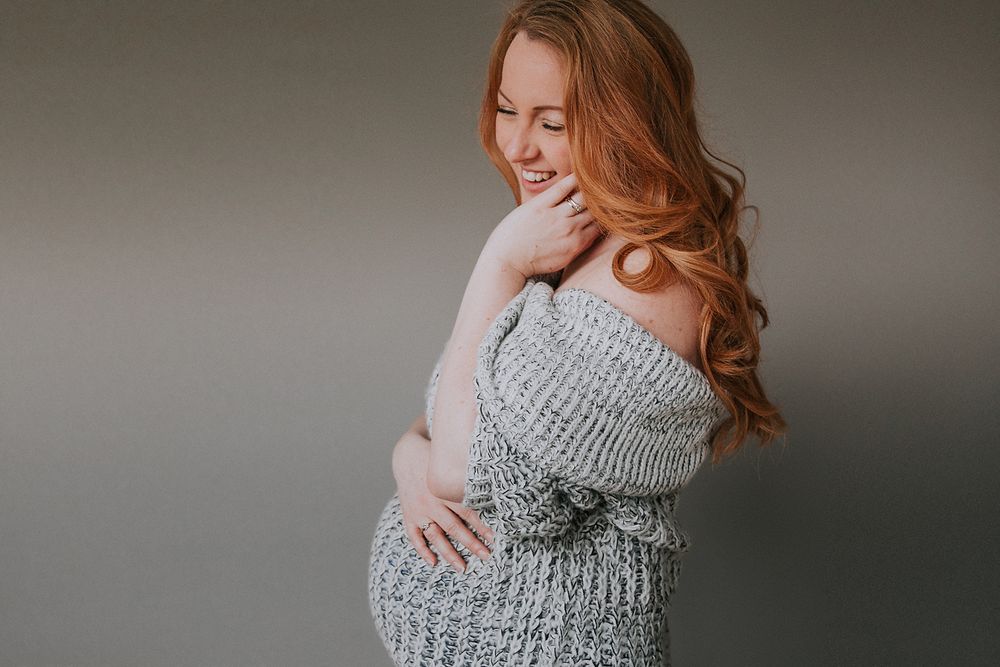 Woman in gray knit sweater smiles warmly while embracing herself against dark backdrop.