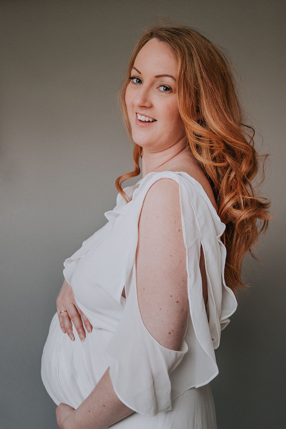 Woman with flowing red hair poses in white ruffled top against gray backdrop in romantic boudoir photoshoot series.