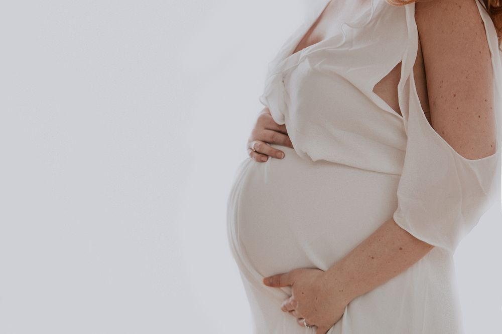 Side view of a pregnant woman in a white dress holding her belly against a light background.