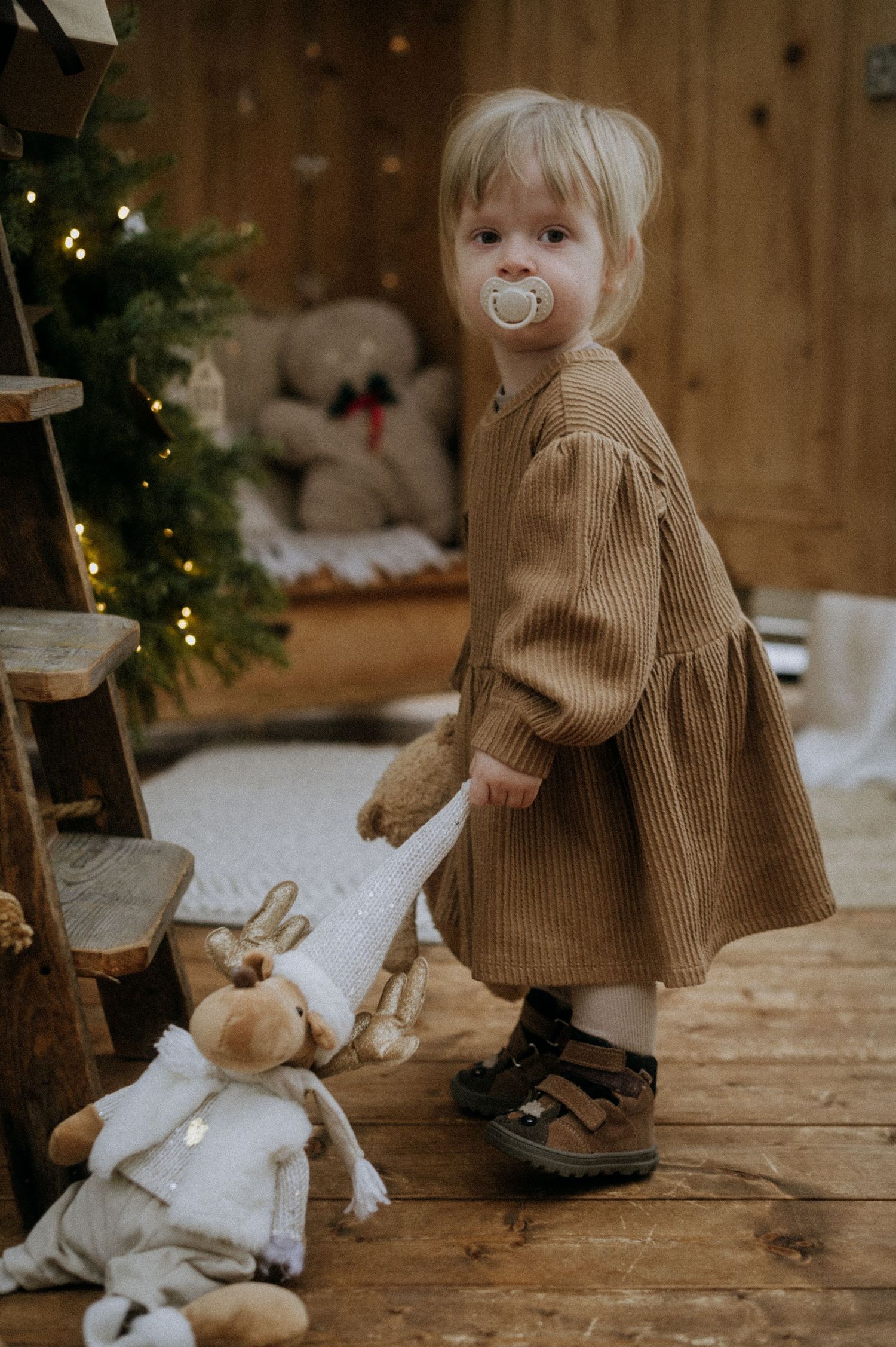 A small child in a brown sweater stands near a Christmas tree with stuffed animals on a rustic wooden floor.