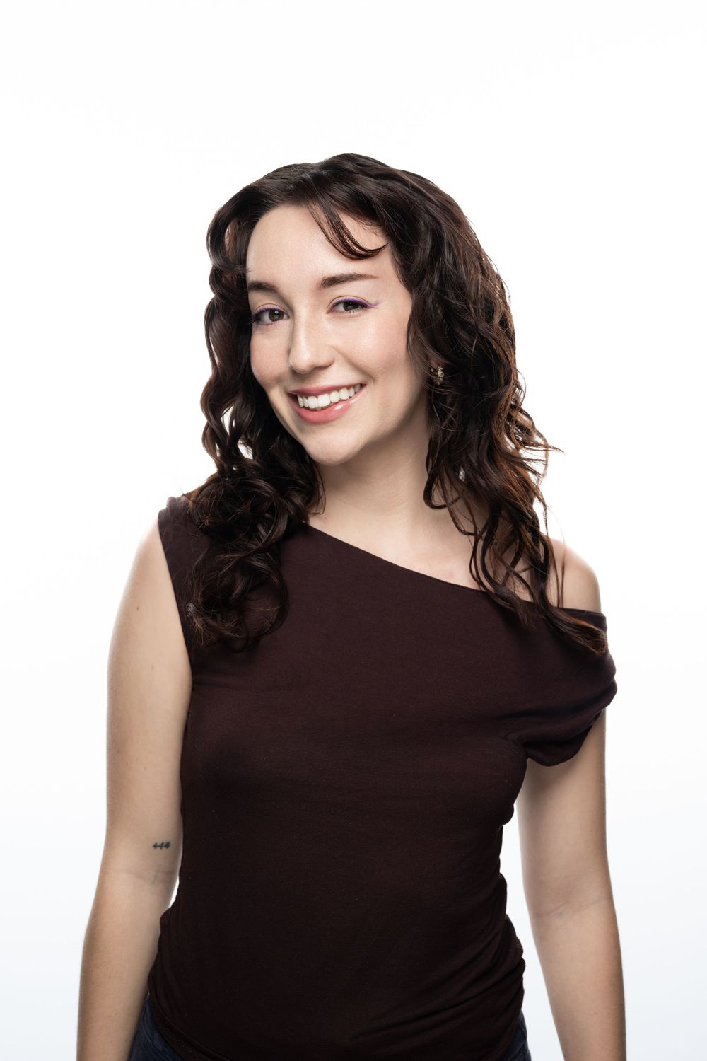Young woman with curly brown hair smiling confidently, wearing a brown asymmetric top against white background.