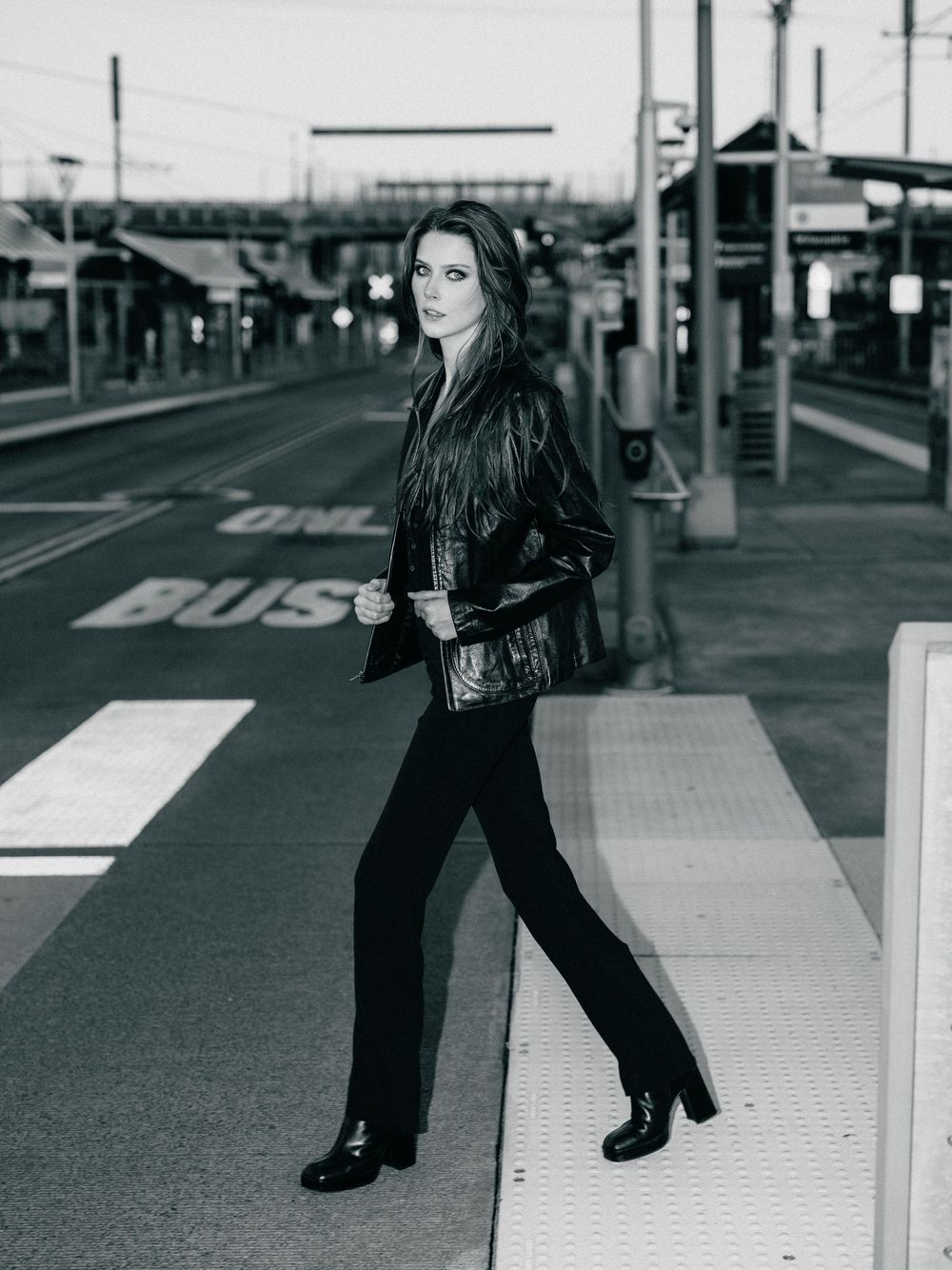 Woman in black jacket and flared pants poses at a bus station crosswalk in a black and white photo.