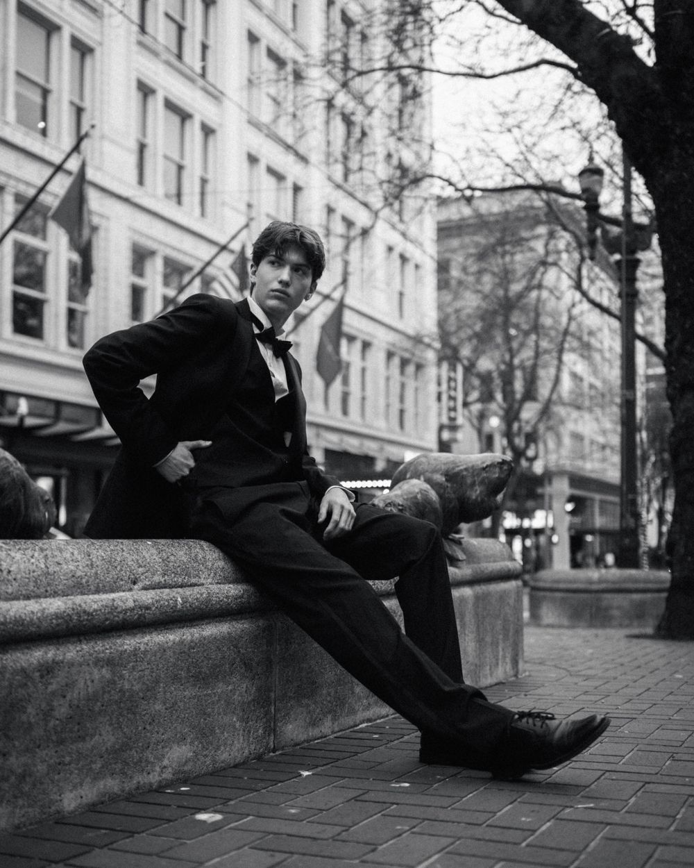Young man in formal black suit sitting on urban stone bench, black and white city street photography.