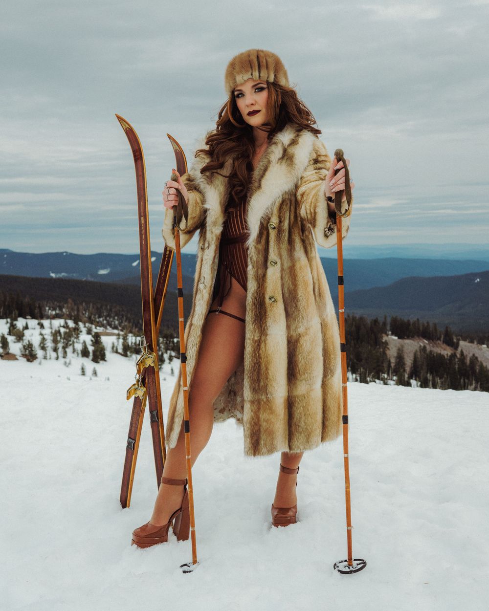 Woman in fur coat and hat holding vintage wooden skis on snowy mountain summit with forest backdrop.