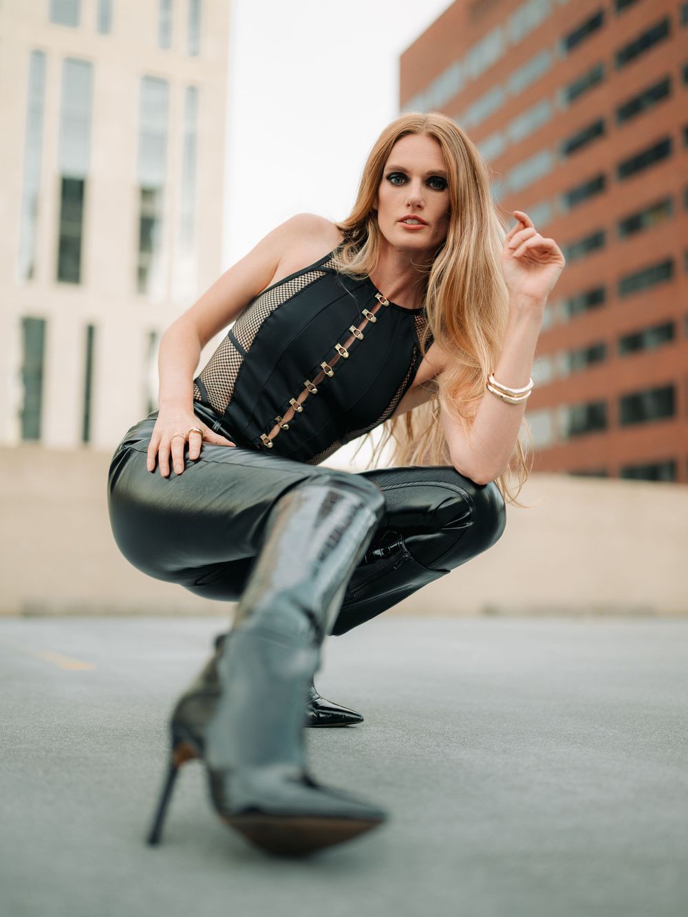 Model in black leather outfit and knee-high boots crouches in urban setting with brick buildings in background.