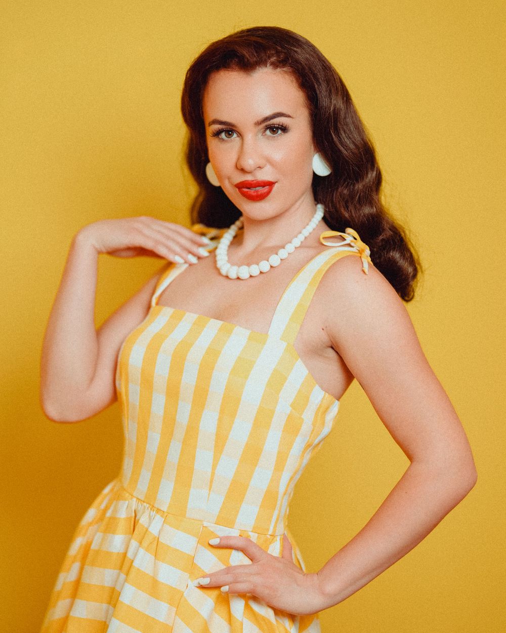 A vintage-style photo of someone in a yellow and white striped sundress and pearl necklace against a yellow backdrop.