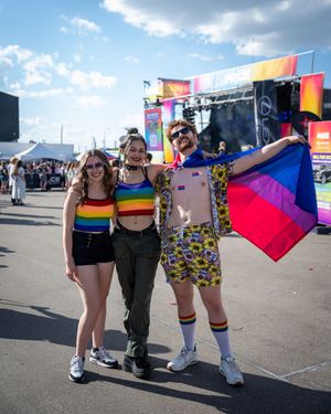 Pride festival attendees wearing colorful outfits and rainbow accessories gather at an outdoor event under cloudy skies.