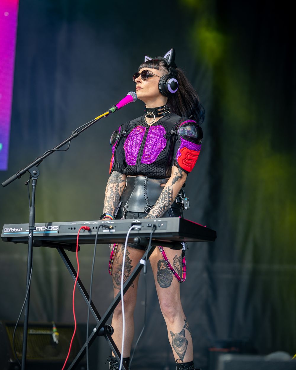 Musician in black leather and pink outfit performs on stage with electronic equipment and microphone setup.