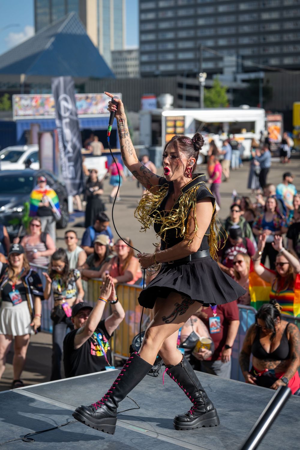 Energetic performer in black dress and combat boots sings on outdoor stage at pride festival with rainbow flags visible.