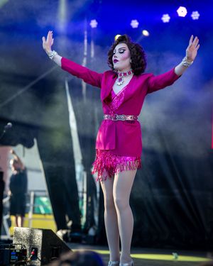 Performer in bright pink fringed dress and silver belt strikes dramatic pose on stage with arms raised during show.