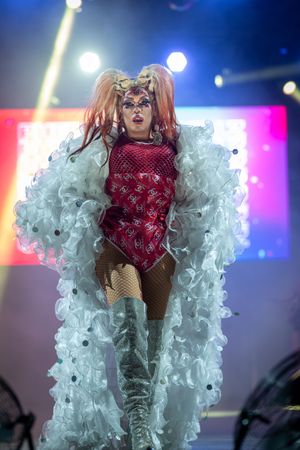 Performer in red bodysuit and white ruffled costume dances on stage with colorful lighting effects in background.