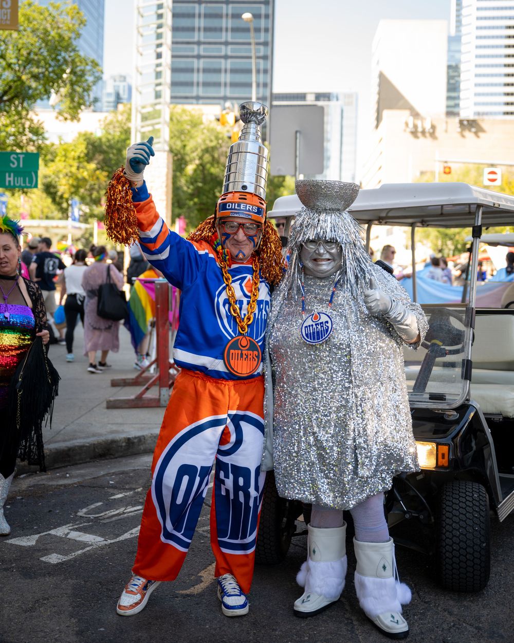 Two mascots in sports team attire, one in blue and white and one in silver sparkly costume, pose together at an outdoor event.