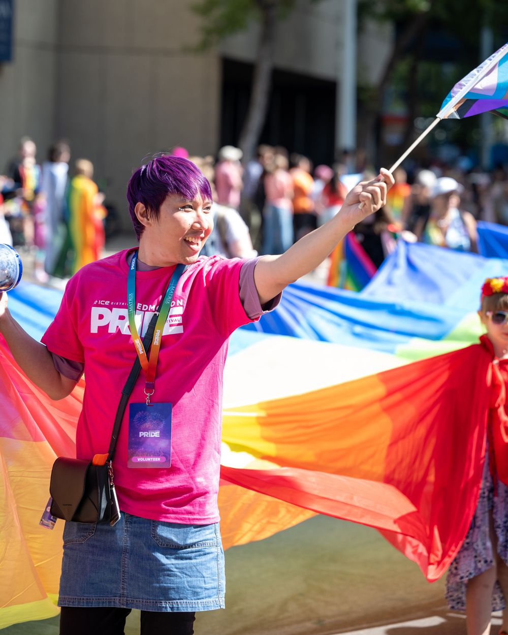 Pride participant in pink shirt waves flag at parade with rainbow banner visible in background at LGBTQ celebration.
