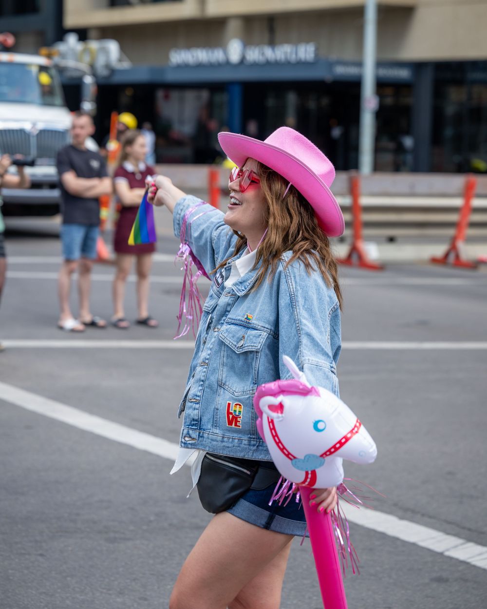 A person wearing denim shorts and a pink hat dances on a city street with an inflatable unicorn pool float.