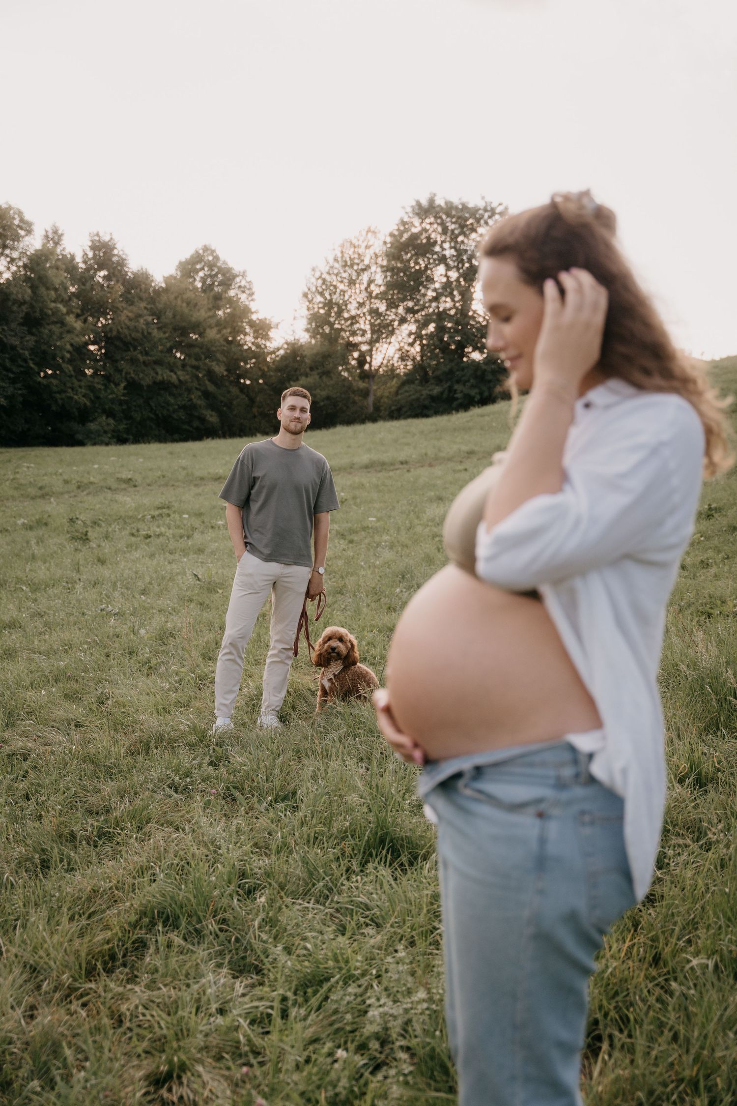A couple and their goldendoodle dog pose together in a sunny grassy field at sunset.