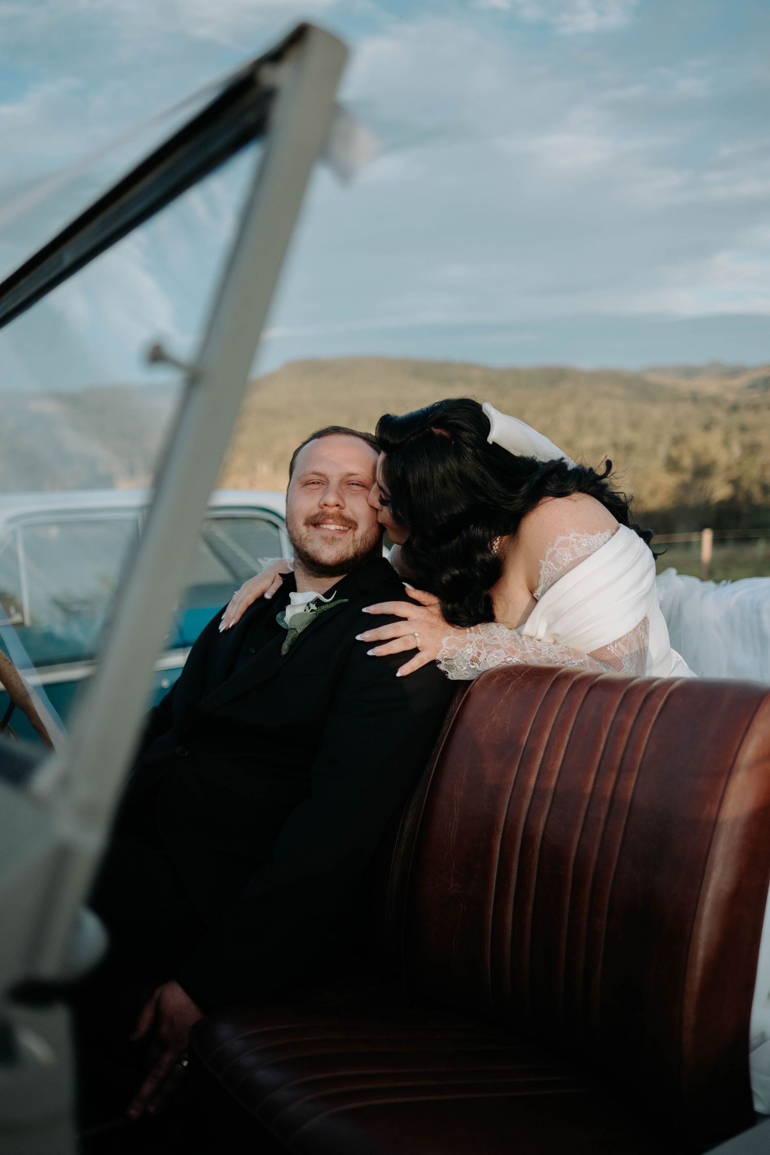 A couple shares an intimate moment in the backseat of a vintage car with brown leather seats against a desert landscape.
