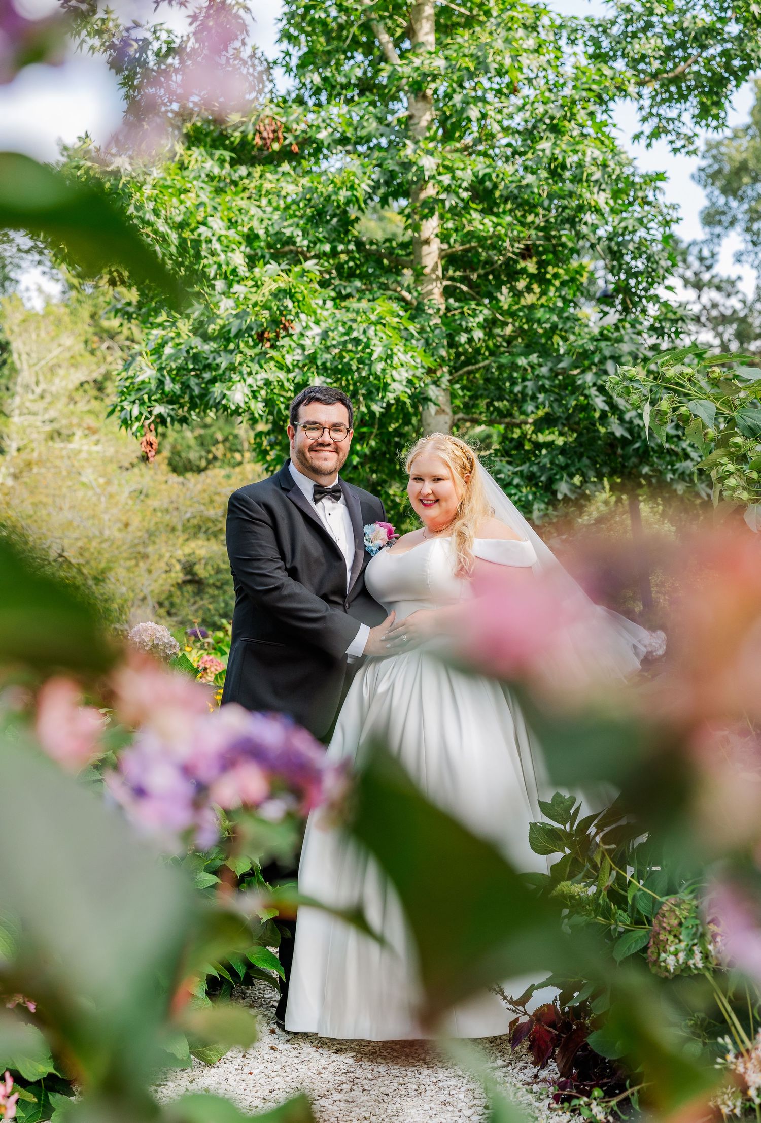 A romantic wedding couple shares a joyful moment together in a lush garden setting with pink flowers in the foreground.