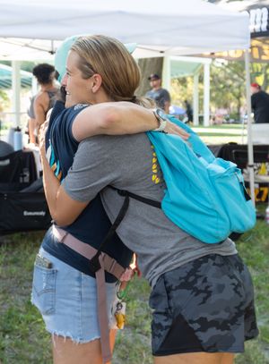 Two people share a warm embrace at an outdoor event with a bright turquoise backpack visible.