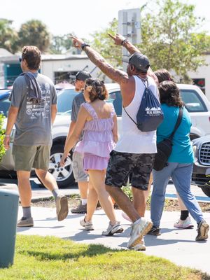 A group of people walking in a line doing silly dance moves together in a parking lot on a sunny day.