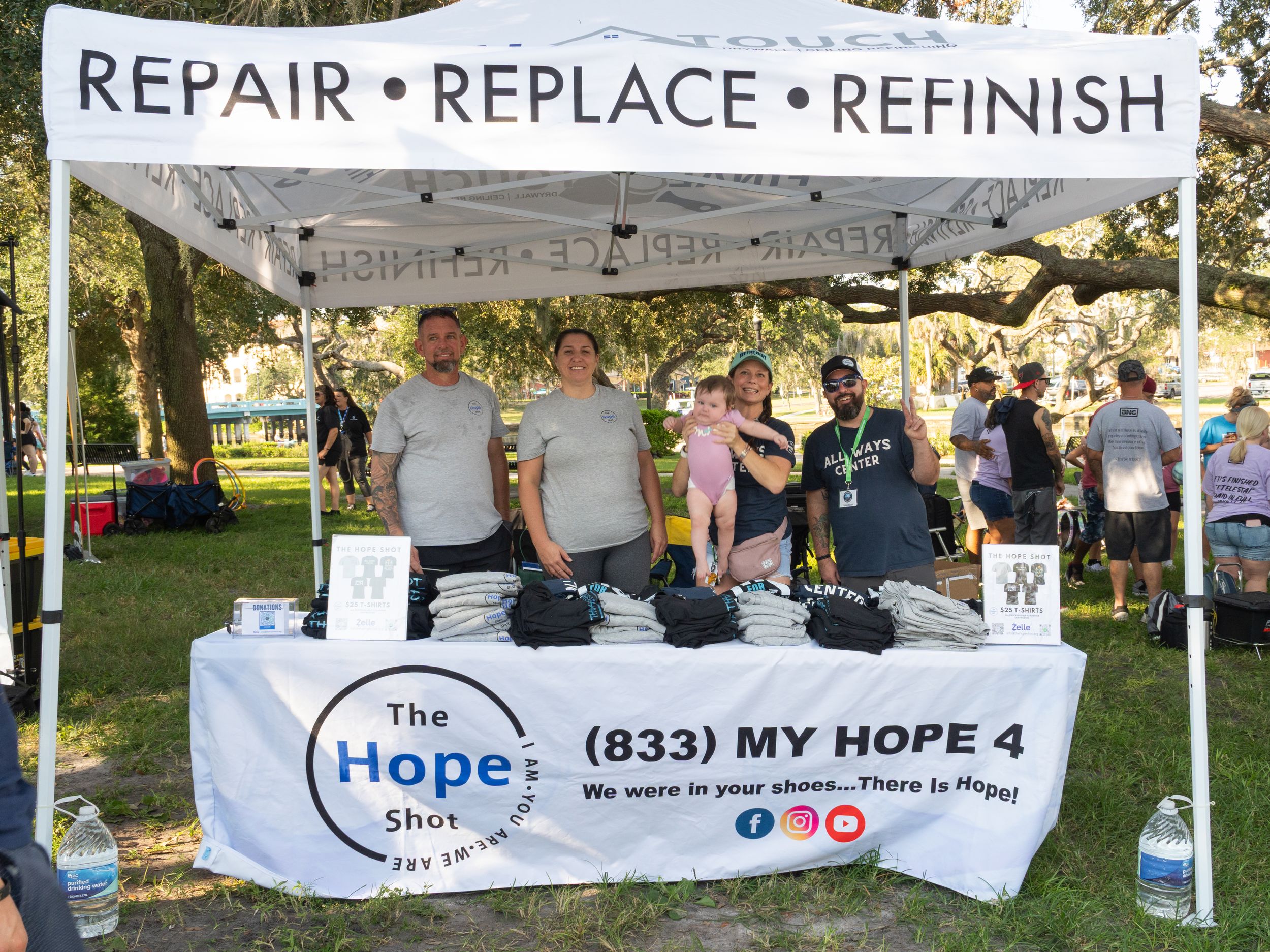 A banner displaying text 'REPAIR REPLACE REFRESH' at an outdoor event with white tents and information booths.