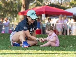 A parent and child sit together on grass during an outdoor event with red tents visible in the background.