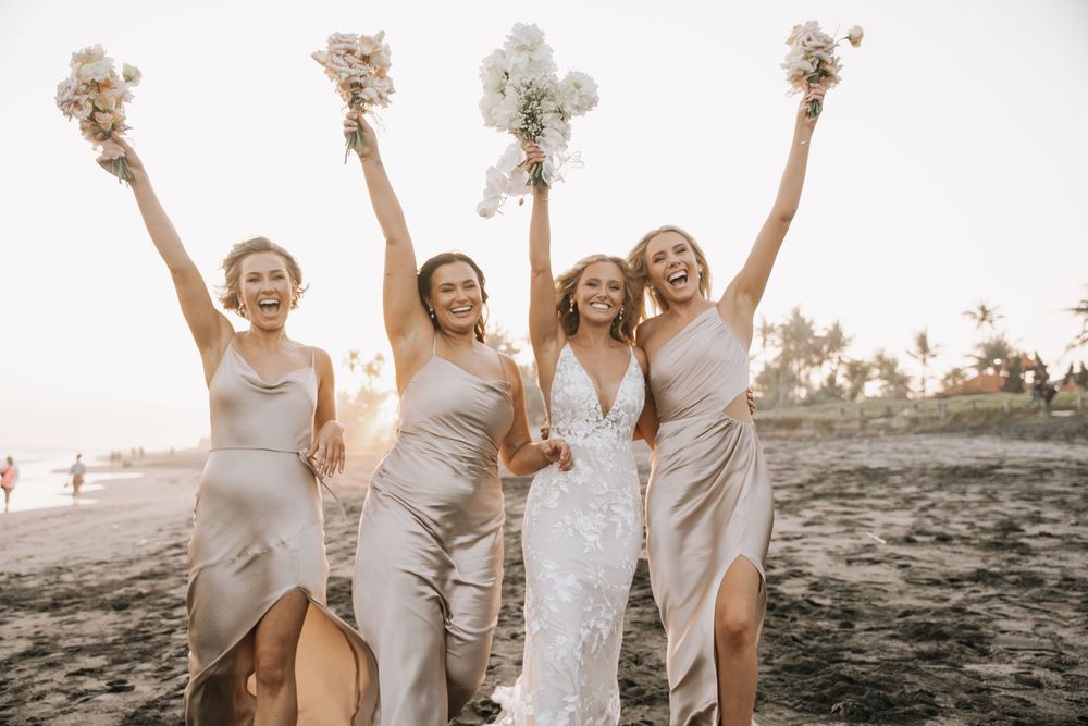 A group in white dresses celebrates joyfully on a beach at sunset, creating a carefree and energetic scene.