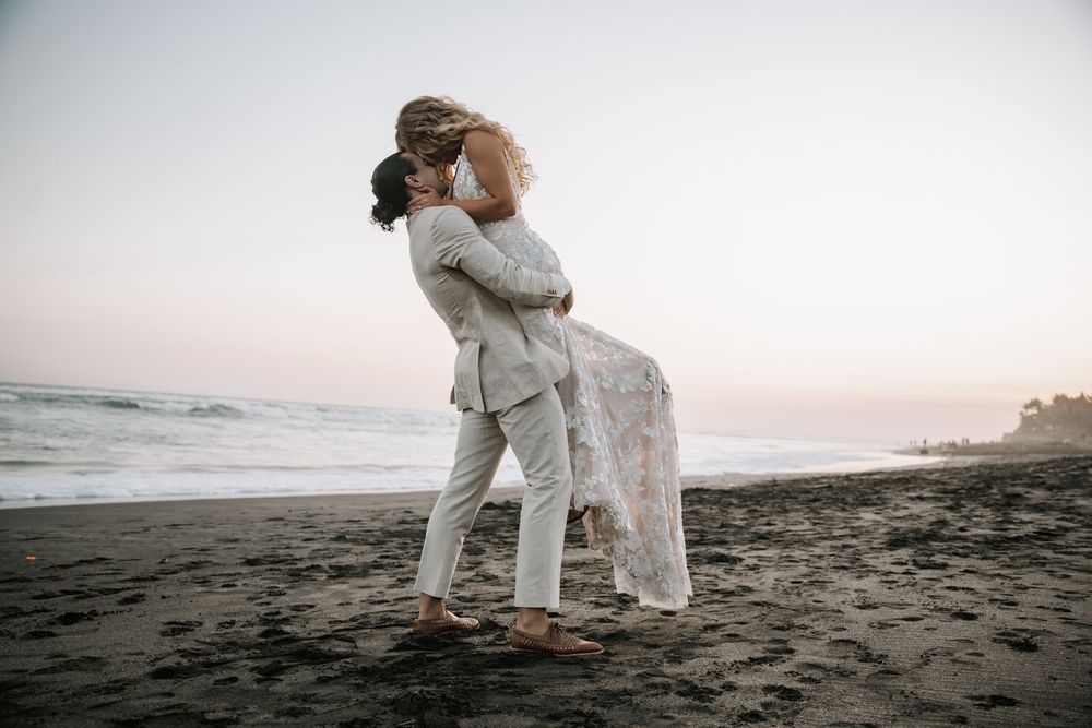 A couple in white outfits embraces on a dark sandy beach at sunset.