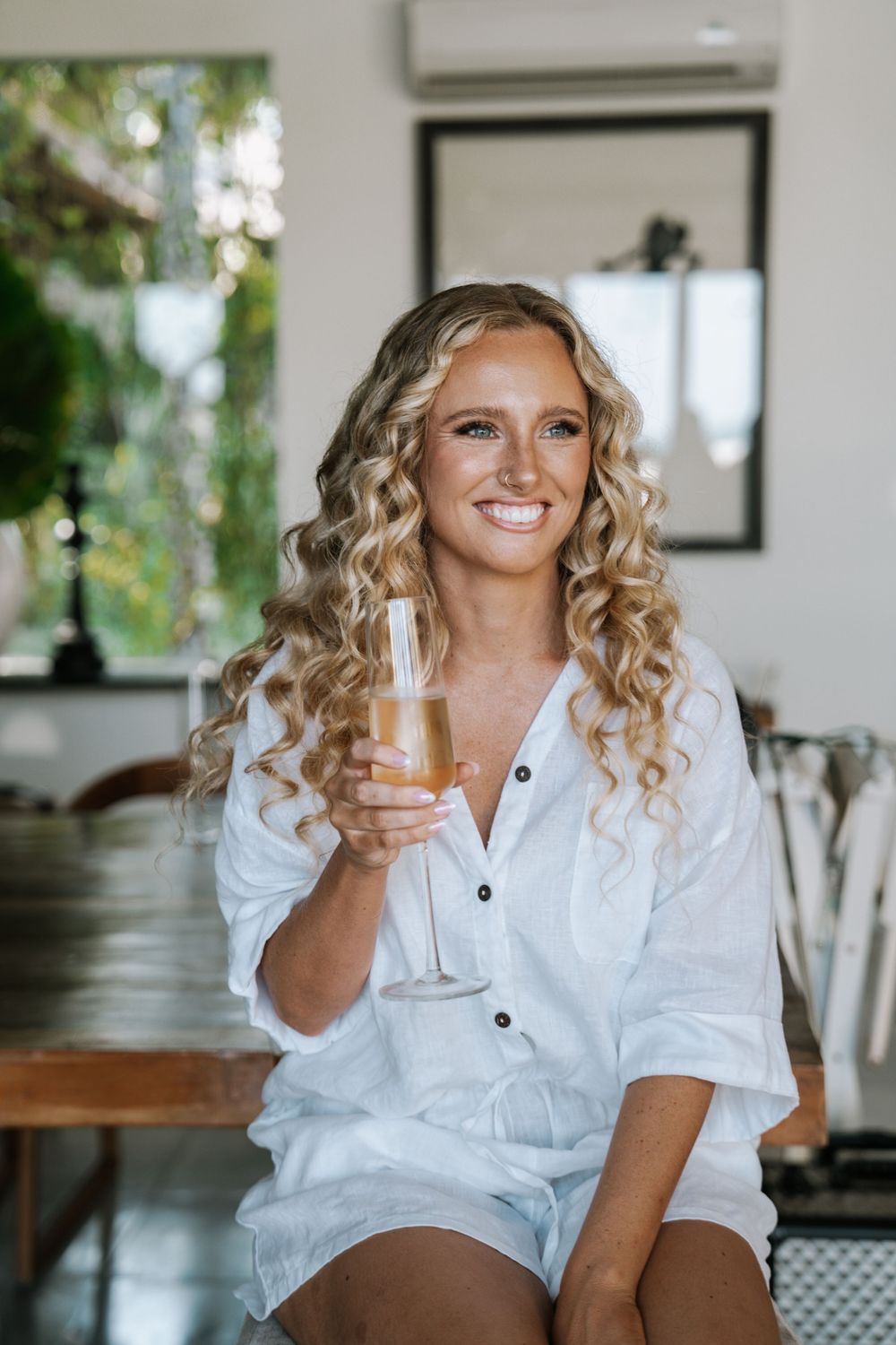 A person in a white button-down shirt holds a champagne glass while sitting and smiling in a bright interior setting.
