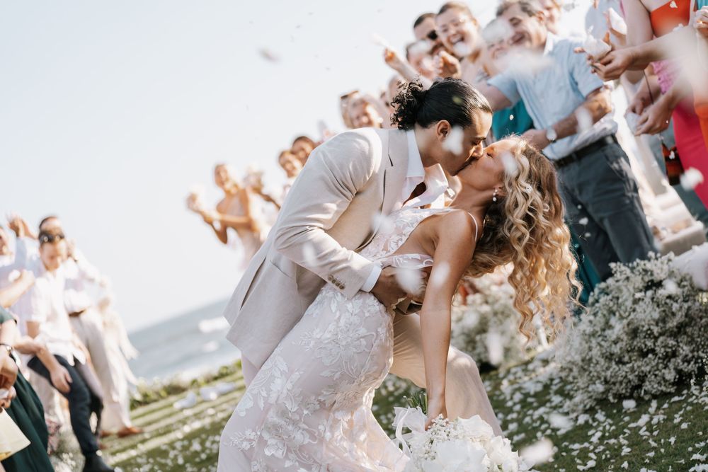 A romantic couple shares a tender kiss during their outdoor wedding ceremony surrounded by guests and blooming flowers.