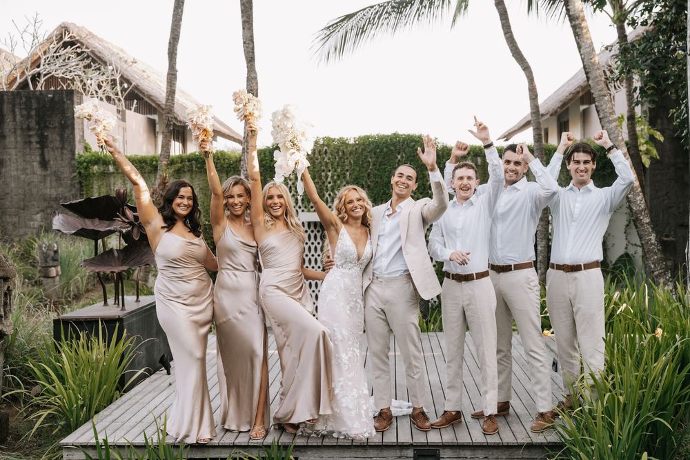 Wedding party celebrates joyfully in a tropical garden setting wearing coordinated champagne and white attire.