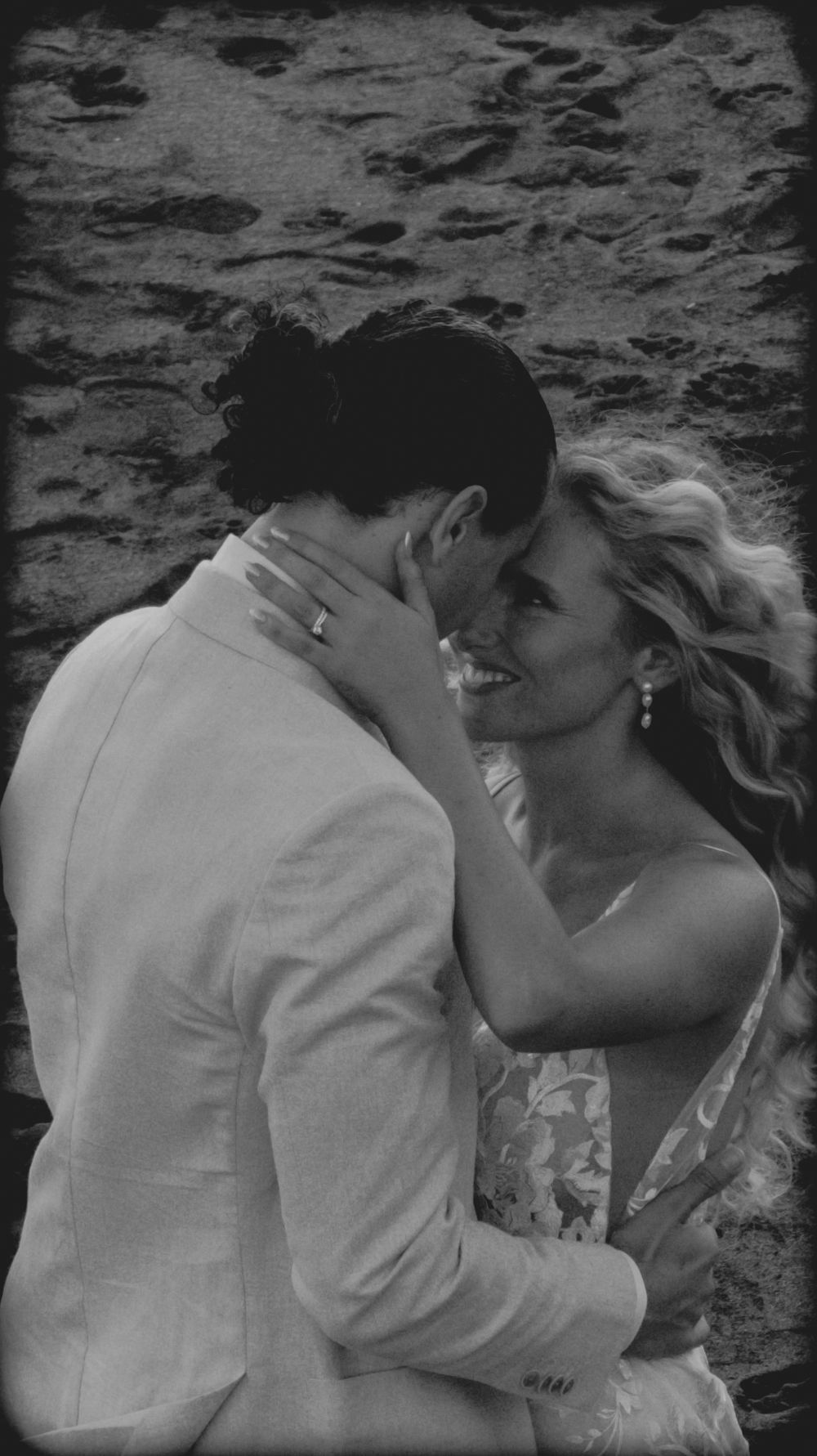 A romantic black and white wedding photo showing a tender moment between a couple embracing against a textured wall.