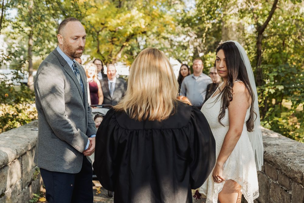 A wedding party gathers outdoors on a sunny autumn day with golden foliage in the background.