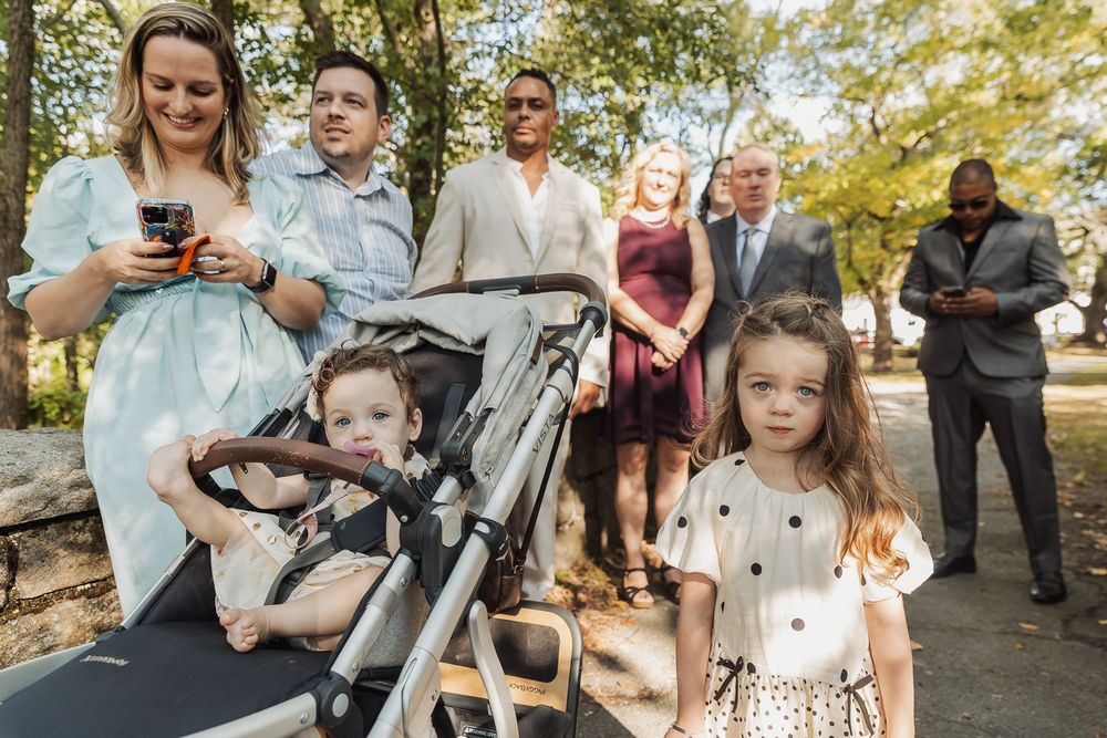 Group of people gathered outdoors on a path with a stroller, dressed in formal attire for a special occasion.