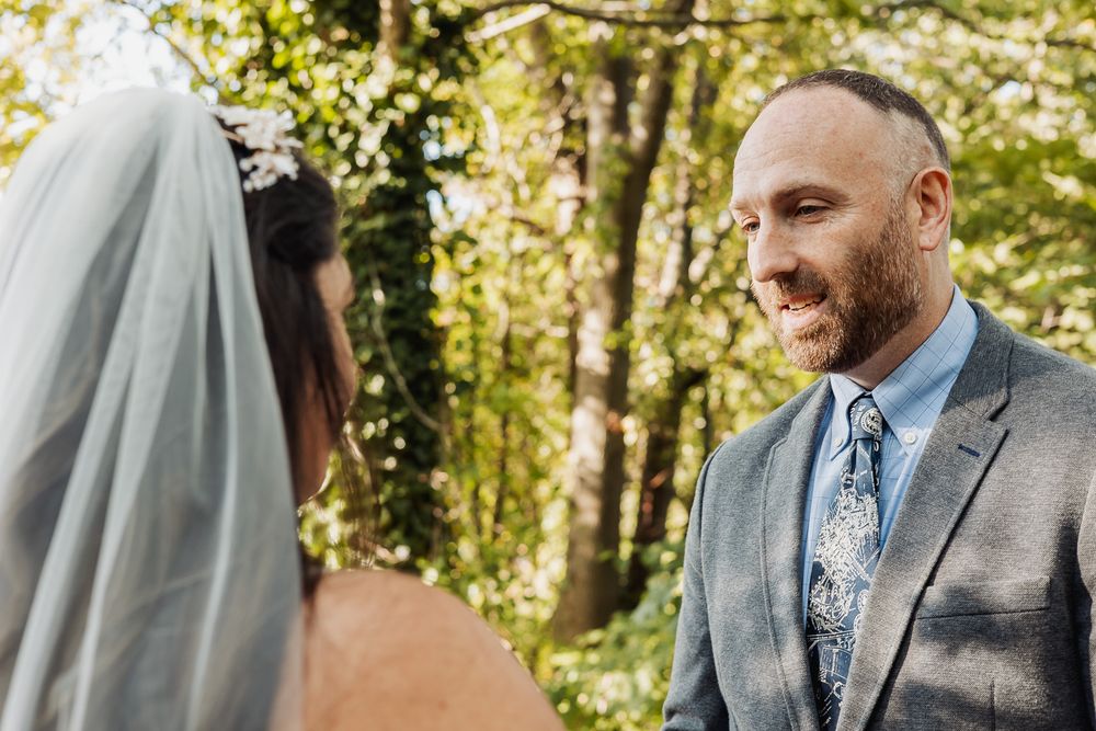 A couple in wedding attire shares a tender moment in a sunlit forest setting during their outdoor ceremony.