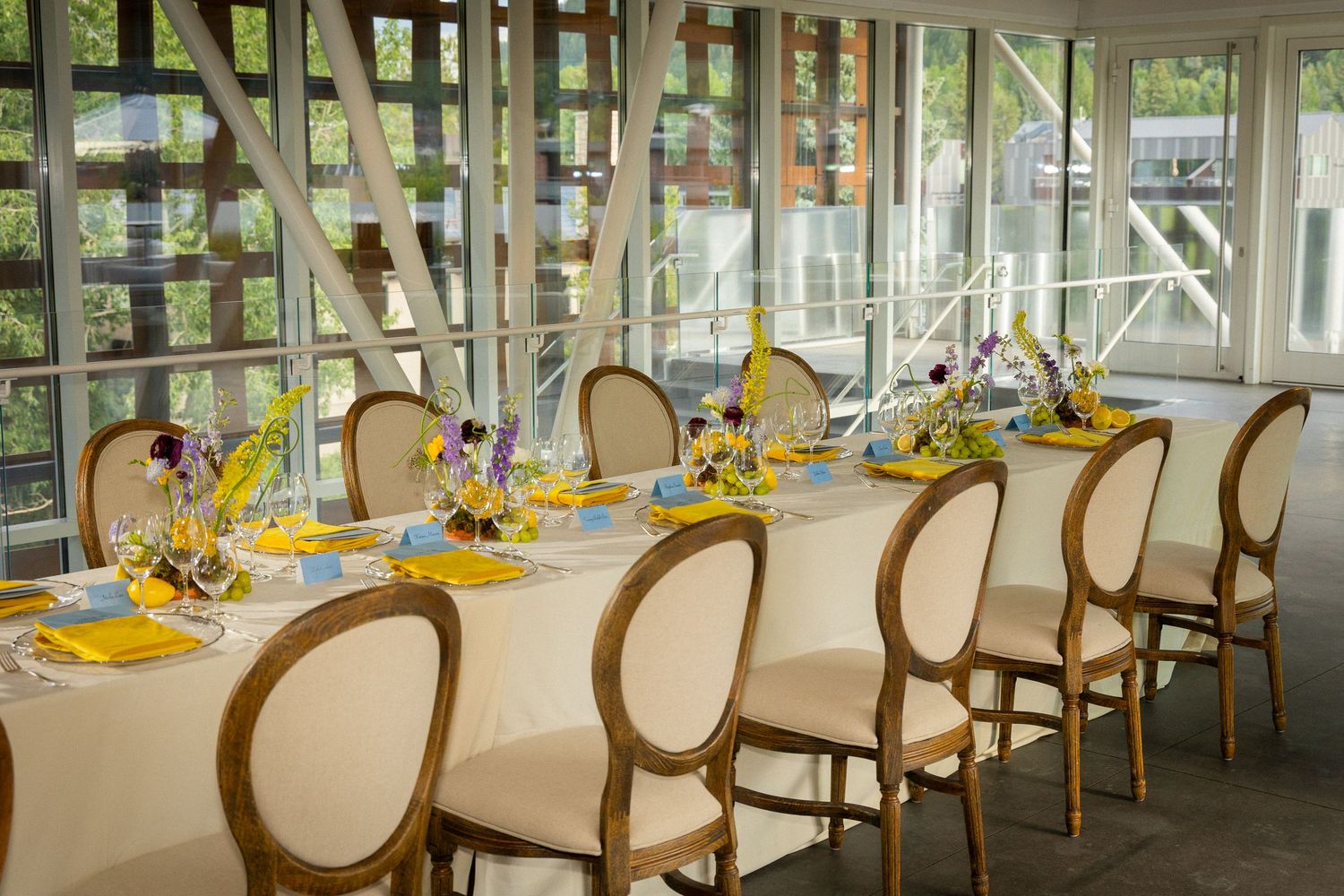 An elegant banquet hall with white chairs and yellow table settings arranged in rows by large windows overlooking greenery.