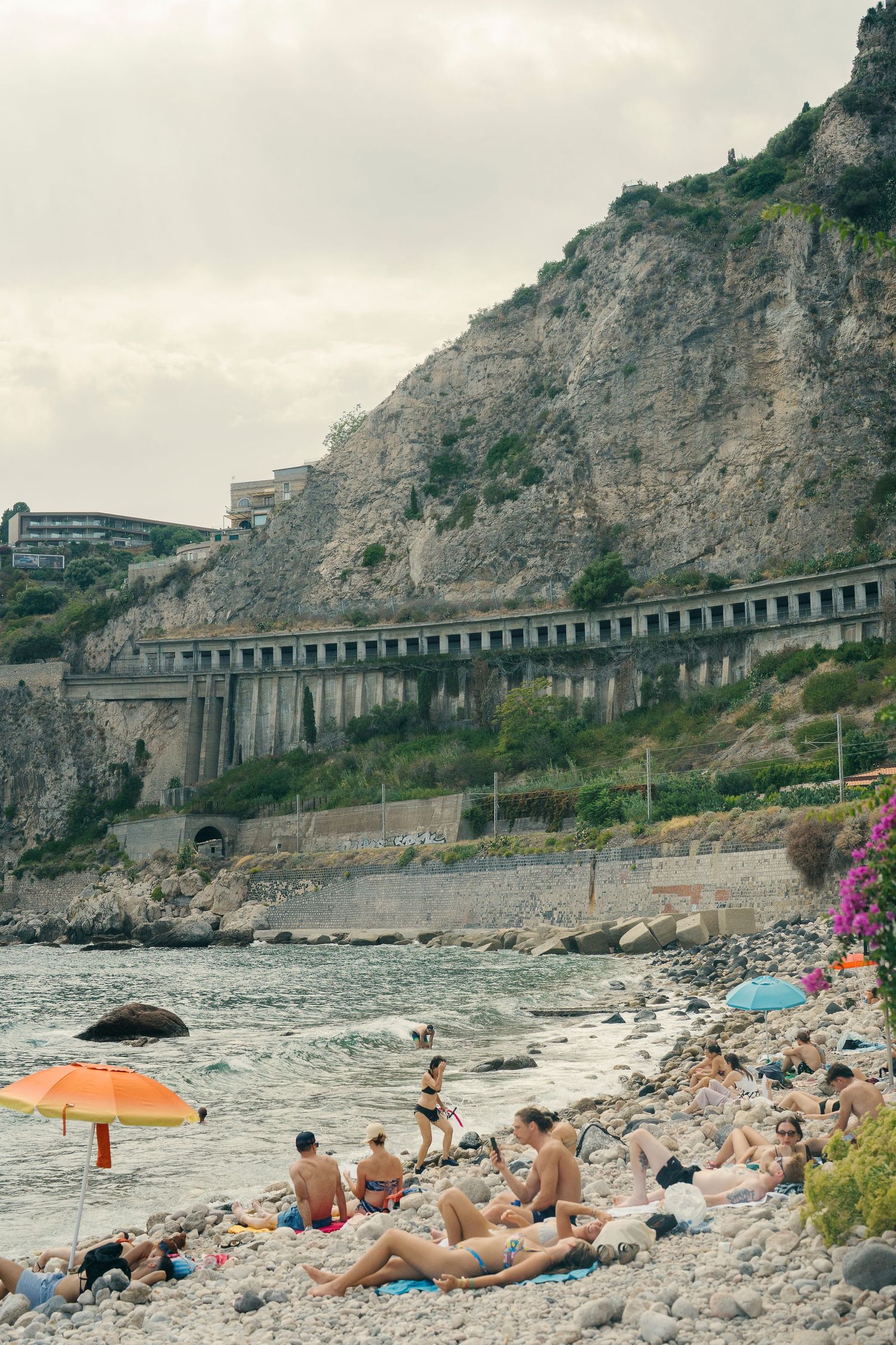 Beachgoers relax on rocky Mediterranean shore beneath dramatic coastal cliffs and railway viaduct in Italy.