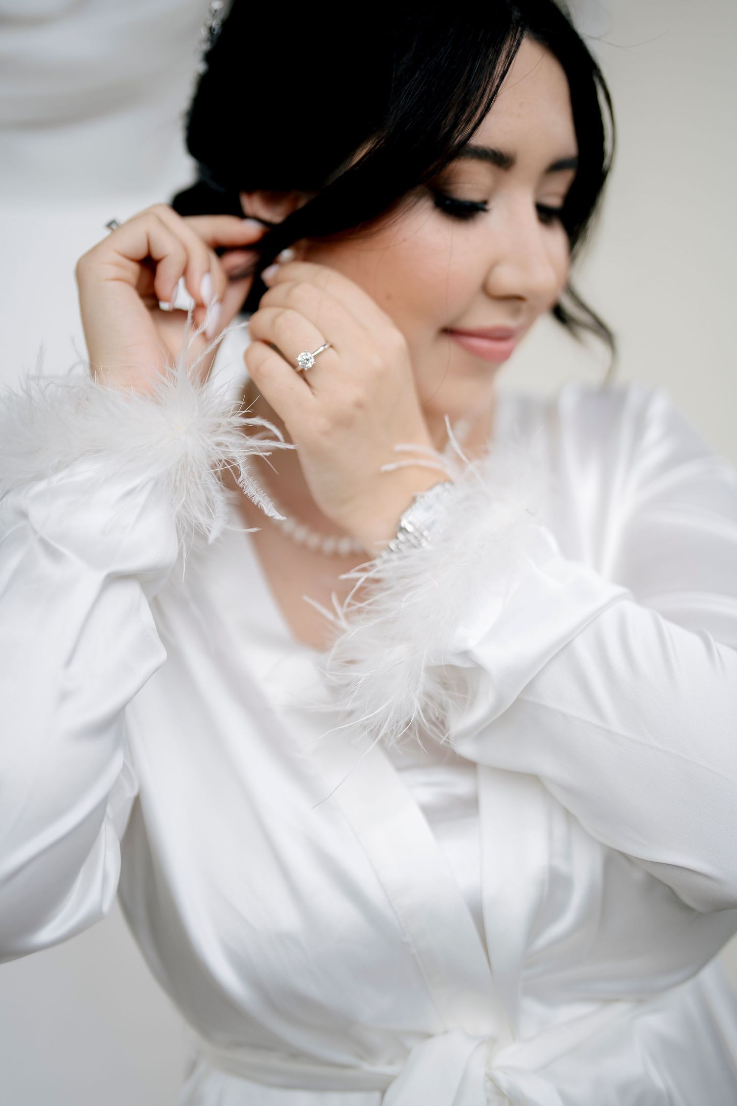 Woman in white satin robe with feather trim poses elegantly against light background in bridal boudoir photoshoot.