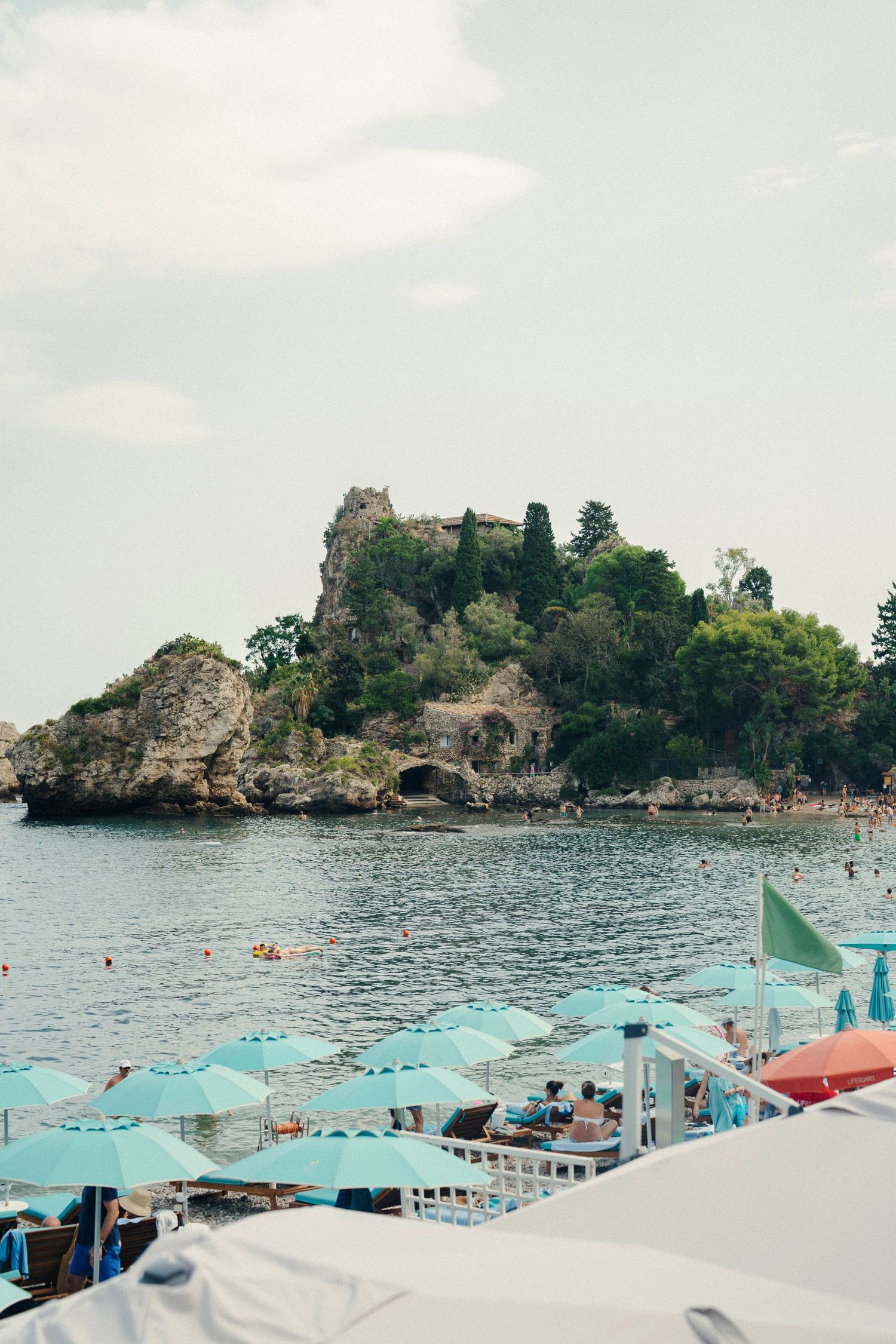 Panoramic view of a rocky Mediterranean coastline with turquoise beach umbrellas and calm waters in summer.