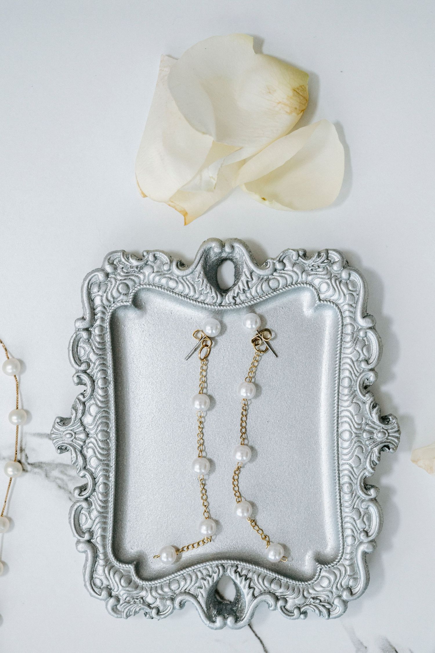 Delicate pearl drop earrings displayed on an ornate vintage silver frame with white rose petals.