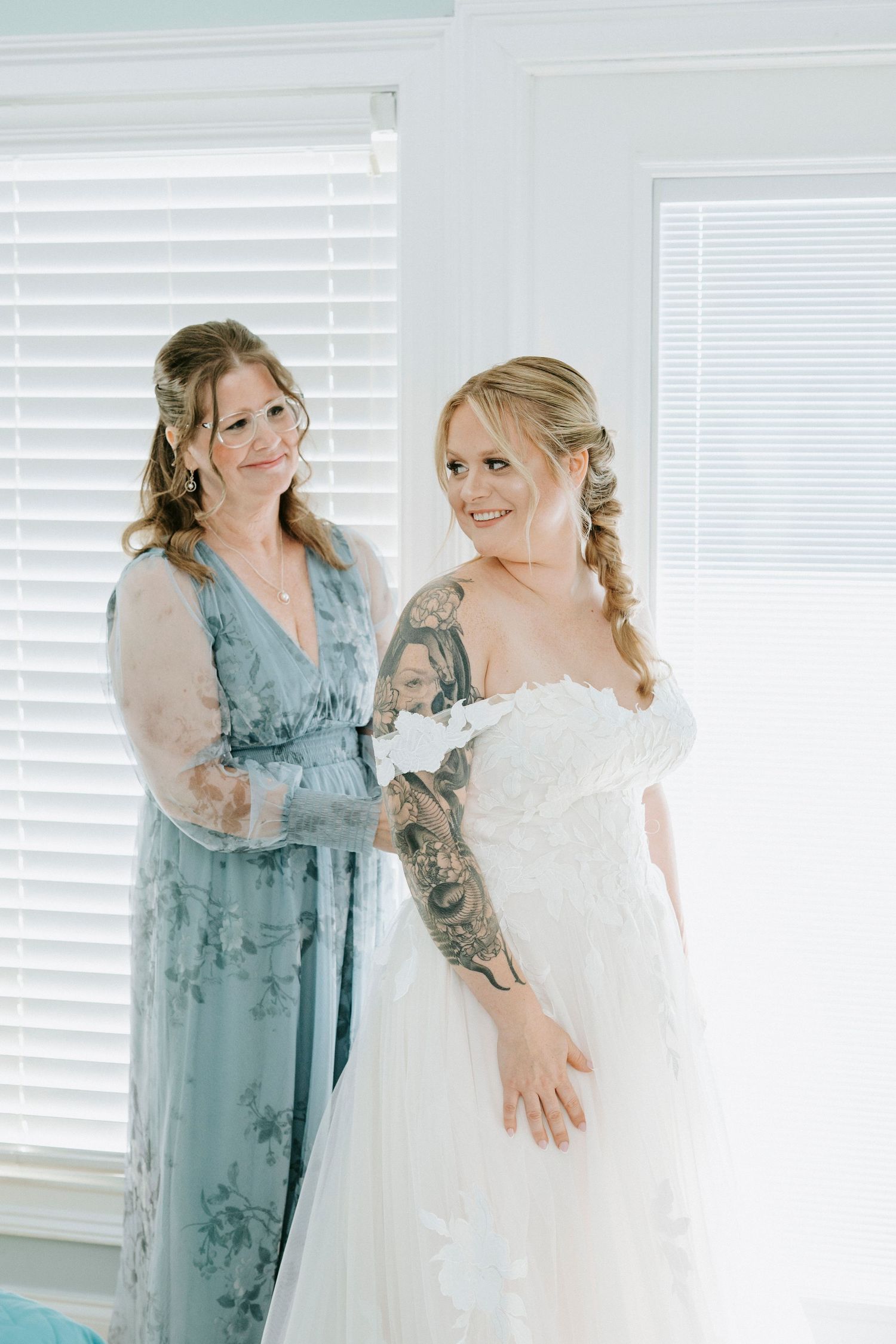 Two women in formal attire share a joyful moment by a window with white blinds.