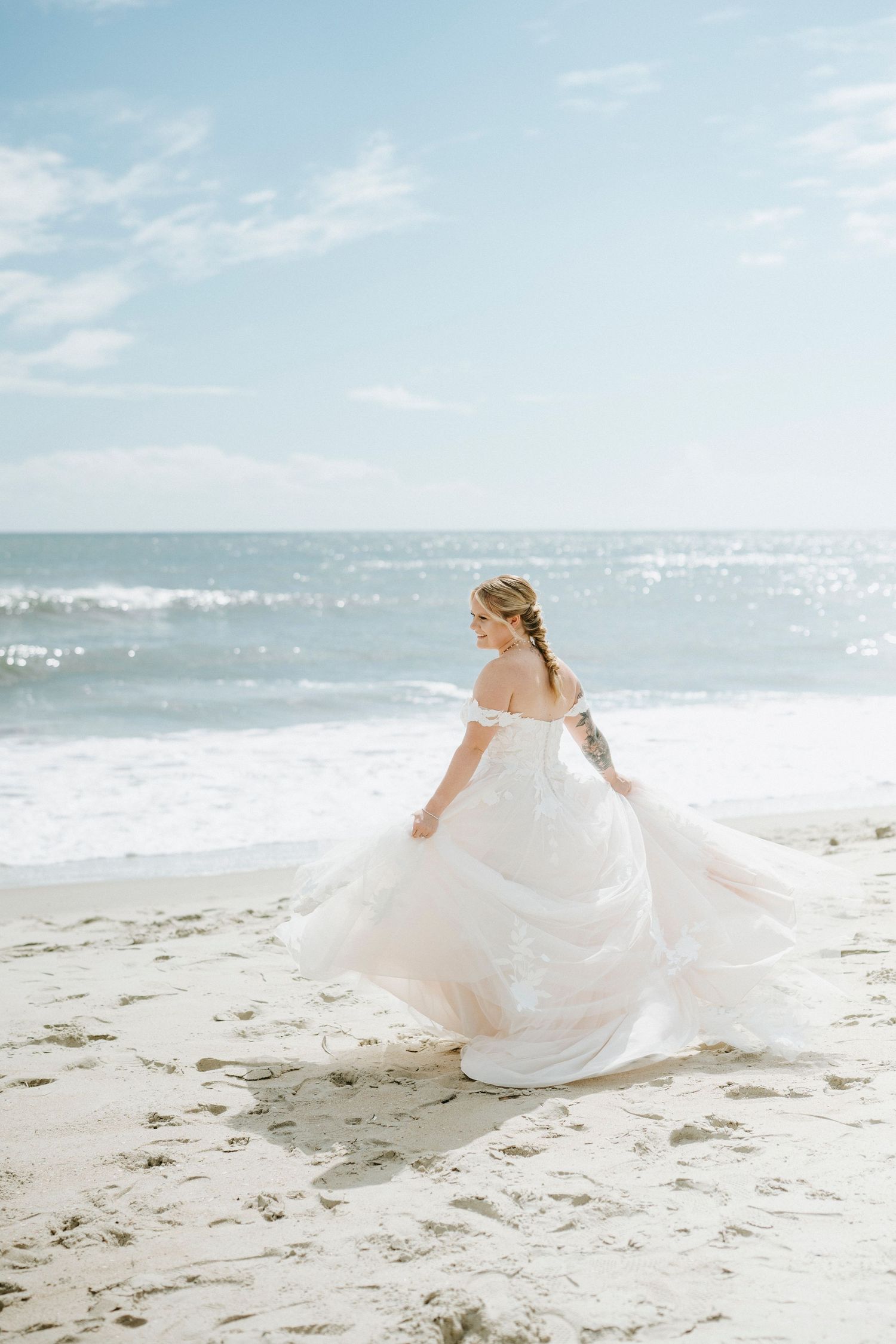 A flowing white wedding dress billows in the ocean breeze as someone twirls on a sunny beach.