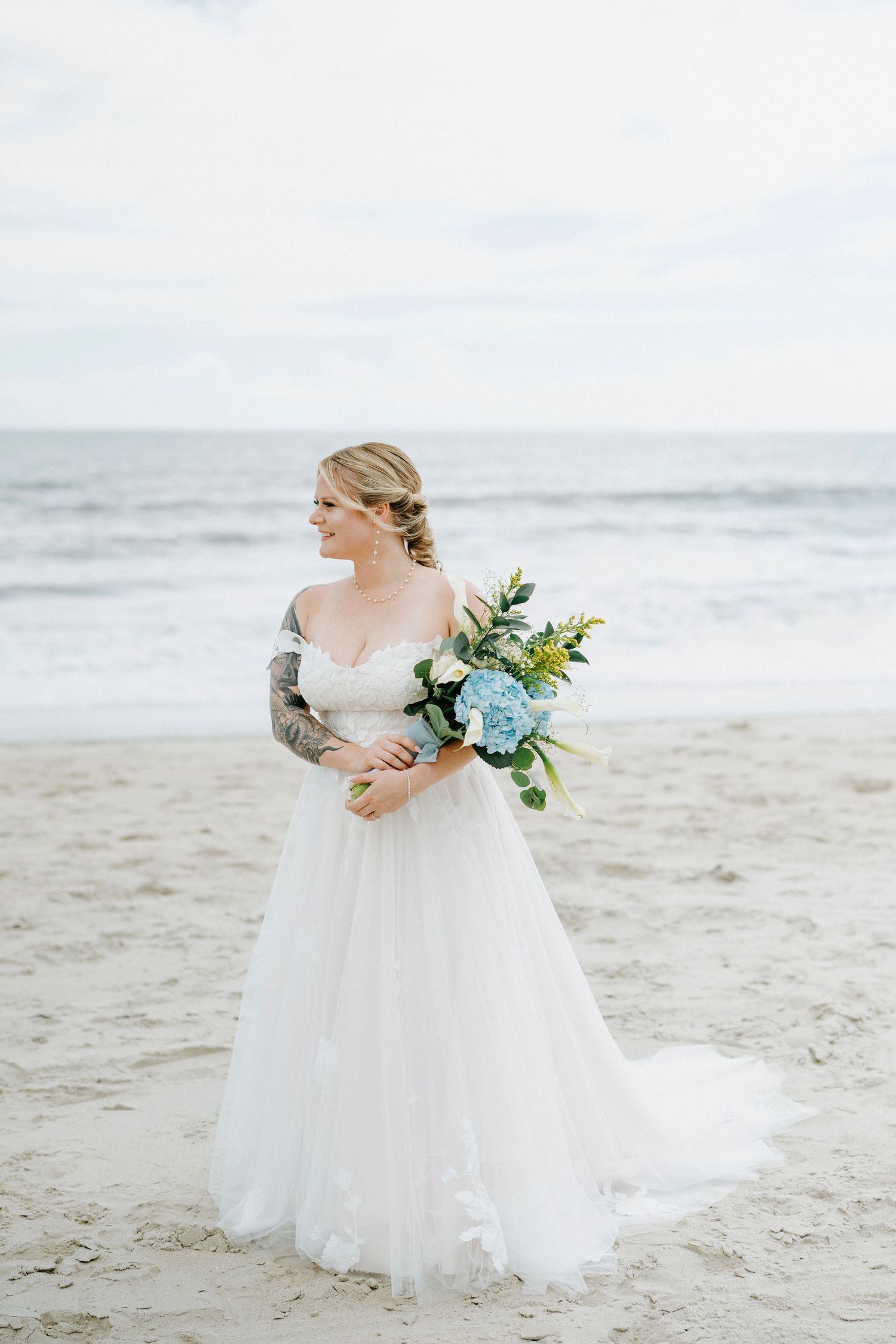 A bride in a flowing white dress holds a blue and yellow bouquet while walking along a serene beach.