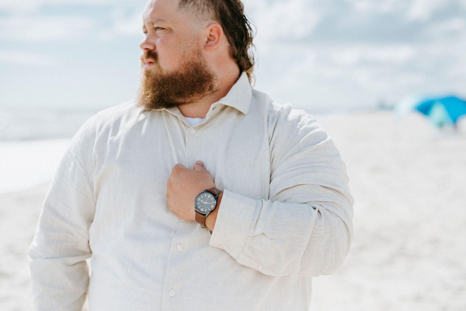 A person in a white button-down shirt stands on a beach wearing a wristwatch against a bright sky.