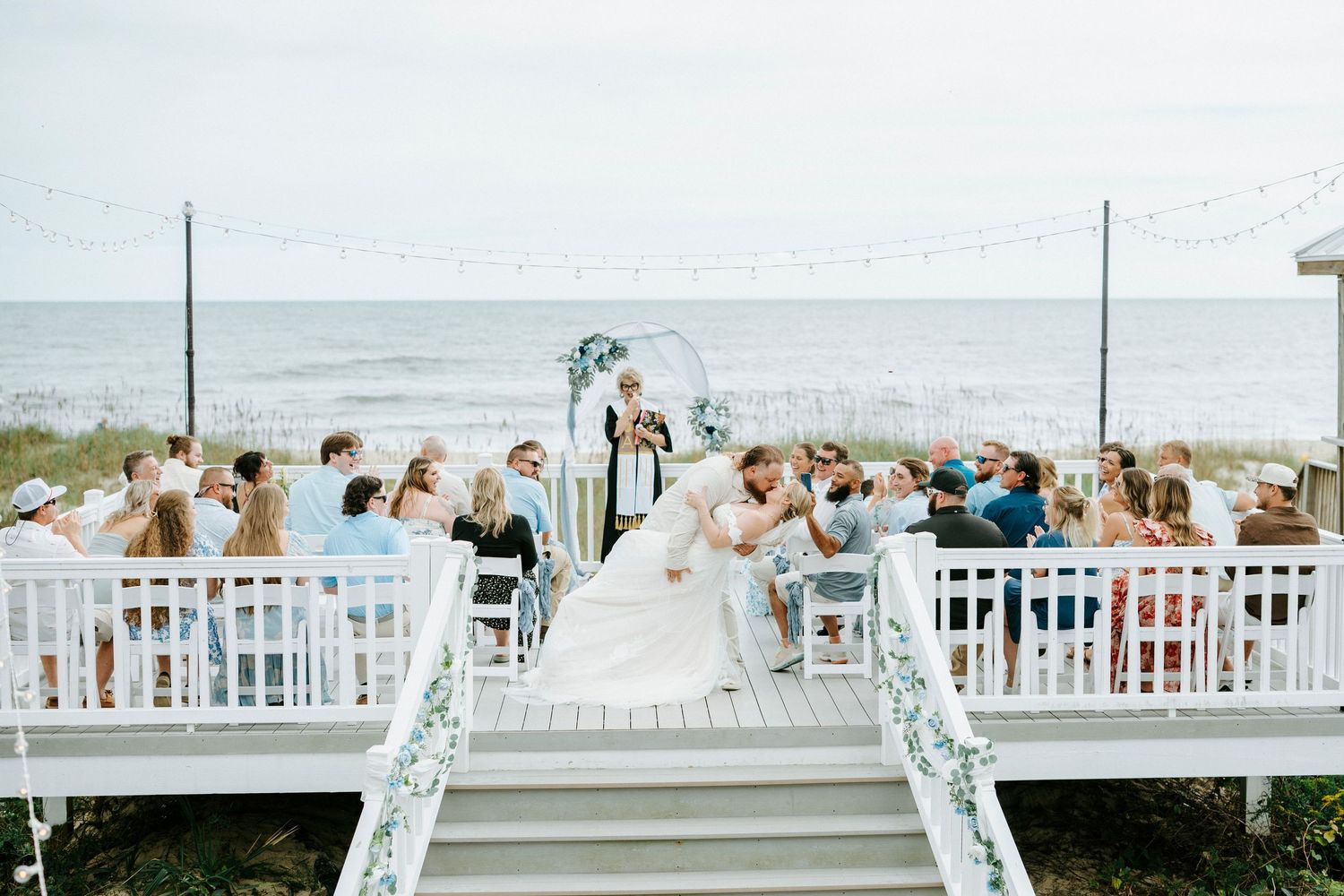 Wedding ceremony guests seated in white chairs overlooking a scenic beach view at an outdoor venue.