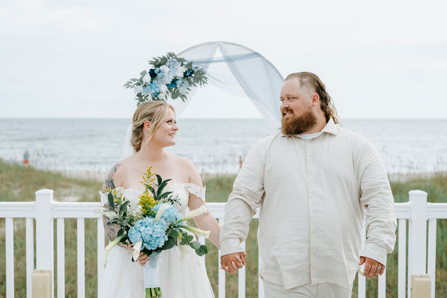 A couple shares a romantic moment on a beachfront deck wearing light colored attire and blue floral accents.